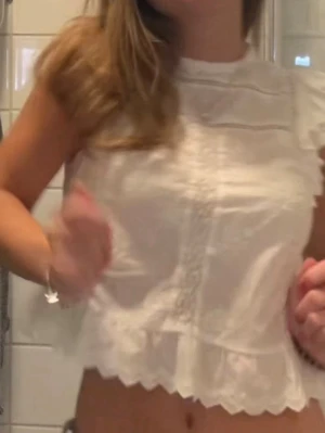 Vit broderad blus med volang - Söt vit blus med broderade detaljer och volangkant. Ärmlös modell med rund halsringning och spetsmönster längs kanterna. Perfekt för sommarens alla tillfällen