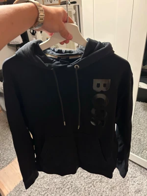 Svart hoodie från BOSS med logga - Snygg svart hoodie från BOSS med stor blank logga vertikalt på bröstet. Hoodien har dragsko i huvan och en klassisk magficka framtill. Tillverkad i mjukt material som känns skönt mot huden. Perfekt för en avslappnad och trendig stil. Storlek M. rätt så tvättad men i acceptabelt skick