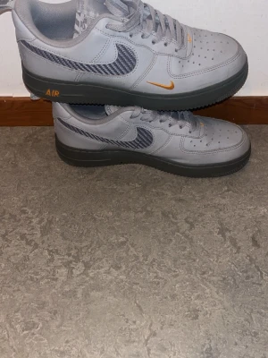 Grå Nike Air Force 1 sneakers - Snygga grå Nike Air Force 1 sneakers med svart och vit rutigt swoosh-märke på sidan. Skorna har mörkgrå sula och orangea detaljer vid loggan och AIR-texten. Tillverkade i läder med klassisk perforerad tå och lågt skaft. Perfekta för dig som gillar streetwear.
