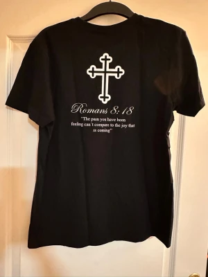 Svart t-shirt med kors och citat - Svart t-shirt från FroccT med vitt kors-tryck på bröstet och större kors samt texten 'Romans 8:18' och citat på ryggen. Klassisk rund hals och korta ärmar. Perfekt för dig som gillar stilrena och statement-plagg.