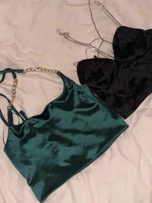 Satin- och chiffongtoppar i olika färger - Tre trendiga toppar: en smaragdgrön satintopp med guldfärgad kedja, en svart satintopp med tunna axelband och en röd chiffongtopp med volangdetaljer och asymmetrisk axel. Perfekta för att lyfta din outfit med lyxig känsla och snygga detaljer.
