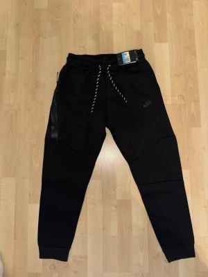 Svarta Nike Tech Fleece mjukisbyxor - Säljer ett par svarta Nike Tech Fleece mjukisbyxor med dragsko i midjan och snörning med vita detaljer. Byxorna har smal passform, ribbade muddar vid benslut och diskret Nike-logga på benet. Snygga detaljer med svarta dragkedjor på sidorna.