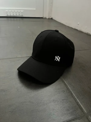 Svart New York Yankees keps - Snygg svart keps från New Era med klassisk böjd skärm och broderad vit NY-logga framtill. Kepsen har justerbar rem bak och är tillverkad i bomull. Perfekt för en sportig och stilren look.