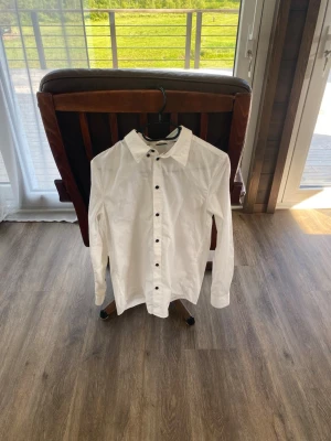 H&M Skjorta - stl 170 - Klassisk vit skjorta från H&M med svarta knappar framtill. Skjortan har lång ärm, normal passform och är tillverkad i mjuk bomull. Perfekt till både jeans och chinos för en clean och stilren look.