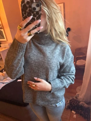 Grå polotröja i stickat - Mysig grå stickad polotröja med hög krage och långa ärmar. Tröjan har en avslappnad passform och ribbstickade muddar vid ärmslut och nederkant. Perfekt för kyliga dagar och enkel att matcha med jeans eller kjol.