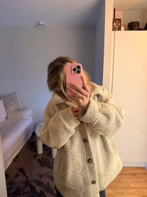 Beige teddyjacka med knappar - Mysig och oversized teddyjacka i beige med stora bruna knappar framtill. Jackan har en klassisk krage och är tillverkad i fluffigt teddy-material som ger en trendig look. Perfekt för kyliga dagar och enkel att styla till olika outfits.