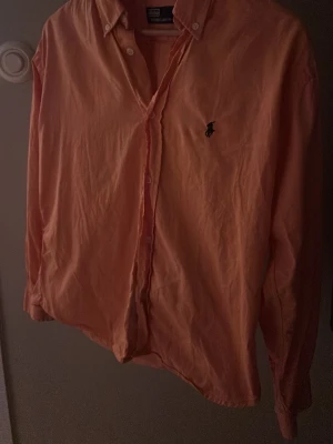 Orange skjorta från Polo Ralph Lauren - Snygg orange skjorta från Polo Ralph Lauren med klassisk krage och knappar framtill. Skjortan har lång ärm och den ikoniska Polo-loggan broderad på bröstet. Perfekt för dig som gillar stilrena och färgstarka plagg. Size L men passar M