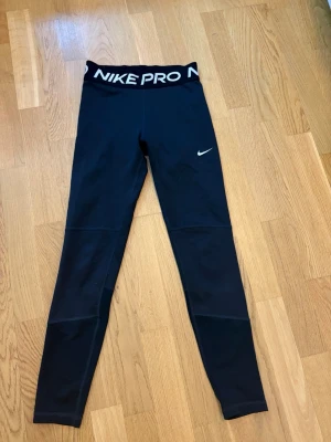 Svarta Nike Pro Dri-FIT tights - Säljer ett par svarta Nike Pro Dri-FIT tights med hög midja och tydlig Nike-logga i midjebandet. Tillverkade i ett stretchigt och fukttransporterande material som passar perfekt till träning. Snygg passform och klassisk design med liten vit swoosh på benet.