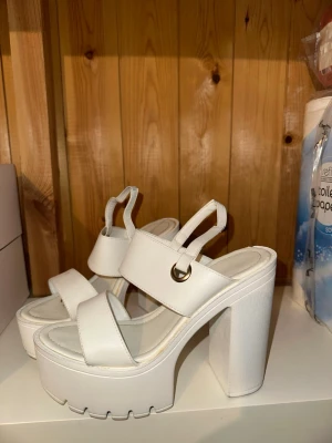 Vita chunky platåsandaler - Säljer ett par vita chunky sandaler med breda remmar och hög platåklack. Sandalerna har en cool 90-talsvibe med grov sula och metallring vid remmen. Tillverkade i syntetmaterial och perfekta för dig som vill sticka ut.