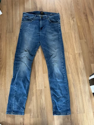 Blå slitna jeans från Lee - Säljer ett par blå jeans från Lee med snyggt slitna detaljer och en liten reva på ena benet. Klassisk femficksmodell med normal midja och raka ben. Jeansen har en tvättad look och är tillverkade i bomull. Stilen LUKE