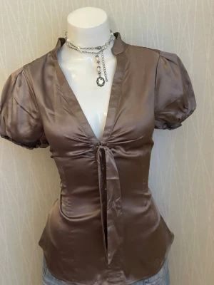 Brun glansig blus med puffärm - Snygg brun blus i glansigt satintyg med puffiga korta ärmar och djup V-ringning. Blusen har en dekorativ knytning framtill och markerad midja för en feminin siluett. Perfekt att styla med jeans eller kjol för en trendig look.