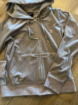 Grå velourhoodie från Juicy Couture - Säljer en grå velourhoodie från Juicy Couture med dragkedja, fickor framtill och broderad JC-logga på bröstet. Hoodien har justerbar huva med guldfärgade detaljer på snörena och ribbade muddar vid ärmslut och nederkant. Perfekt för en chill och trendig look. Märket på dragkedjan är sönder.