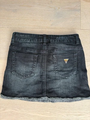 Mörkblå  jeanskjol från Guess - Snygg mötkblå jeanskjol från Guess med slitna detaljer och rå kant nertill. Kjolen har fem fickor, dragkedja och knapp framtill samt en liten guldfärgad Guess-logga på bakfickan. Perfekt till en avslappnad och trendig look.