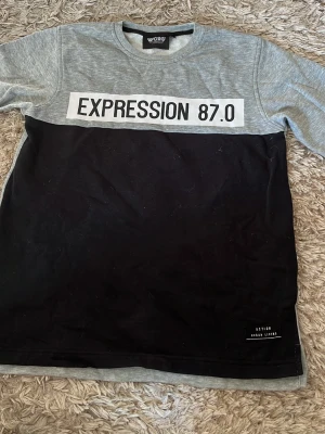Grå och svart t-shirt från Woxo - Snygg t-shirt från Woxo med grå överdel och svarta nedre delen. Tryck med texten 'EXPRESSION 87.0' framtill. Rund halsringning och klassisk passform. Perfekt för dig som gillar streetwear och enkel stil.