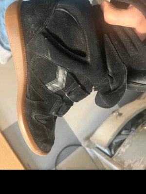 Svarta höga sneakers med kardborre - Säljer ett par svarta höga sneakers med cool design och kardborreband. Skorna har en ovandel i mocka och läderdetaljer samt en klassisk gummisula i brunt. Perfekta för dig som gillar streetstyle och vill ha något unikt på fötterna.