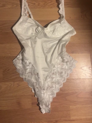 Vit spetsbody  - Supersnygg vit body med spetsdetaljer längs sidorna. Framsidan är i glansigt satinliknande matiral. Köpte för en vecka sen och inte använt den då jag inte tycker jag passar i den men tyvärr glömt kvittot så kan ej lämna tillbaka och därför jag säljer! (Sann i storleken) pris går att diskuteras 