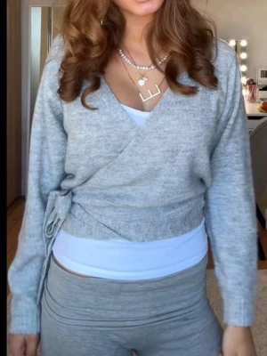 Ljusgrå omlottstickad tröja - Snygg ljusgrå stickad omlott-tröja med knytband i sidan och ribbade muddar. Tröjan har lång ärm och en mjuk, följsam passform som passar perfekt till både jeans och tights. Enkel och stilren design utan synligt märke.