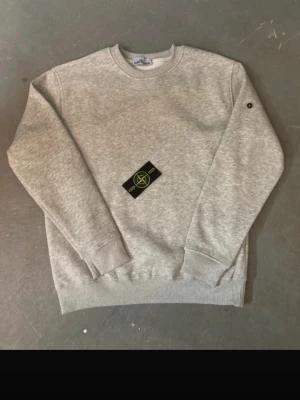 Grå sweatshirt från Stone Island -  Säljer en stilren grå sweatshirt från Stone Island med klassisk rund hals och ribbade muddar. Tröjan har den ikoniska Stone Island-loggan på vänster ärm och är tillverkad i mjukt bomullsmaterial. Perfekt för en avslappnad och trendig look.