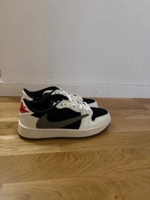 Air Jordan 1 Low Travis Scott svart/vit - Snygga Air Jordan 1 Low Travis Scott sneakers i svart och vitt med grå swoosh och röda broderade detaljer på hälen. Skorna har klassisk låg siluett, läder- och textilmaterial samt ikoniska loggor. Perfekta för dig som vill sticka ut med streetwear-stil.