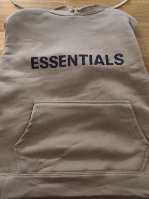 Beige Essentials hoodie med tryck - Snygg beige hoodie från Essentials med svart tryck på bröstet. Klassisk modell med huva, dragsnören och stor känguruficka framtill. Perfekt för en avslappnad och trendig streetwear-look.