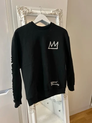 Svart sweatshirt Basquiat print - Cool svart sweatshirt från H&M med Basquiat-inspirerat tryck på ryggen och en vit krona på bröstet. Textdetaljer på ena ärmen och nedtill framtill. Rak passform och rund halsringning. Perfekt för dig som gillar streetwear och konstnärliga detaljer.