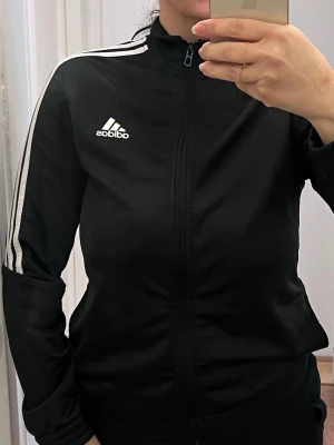 Svart Adidas träningsjacka - Svart träningsjacka från Adidas med klassiska vita ränder längs ärmarna och vit Adidas-logga på bröstet. Jackan har dragkedja framtill, hög krage och är tillverkad i ett glansigt, sportigt material. Perfekt för träning eller chill.