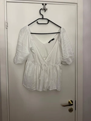 Vit top med puffärm och knyt - Söt vit topp med v-ringning, puffiga korta ärmar och knyt i midjan. Toppen har en luftig passform och är tillverkad i ett mjukt bomullstyg. Perfekt för en fräsch och avslappnad look. Använd sparsamt och inga defekter😊