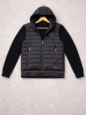 Balmain Cardigan  - Snygg svart pufferjacka från Balmain med huva och dragkedja framtill. Jackan har quiltad kropp, stickade ärmar och ribbade muddar. Två framfickor med dragkedja och Balmain Paris-logga längs nederkanten. Perfekt för kalla dagar och riktigt trendig look.