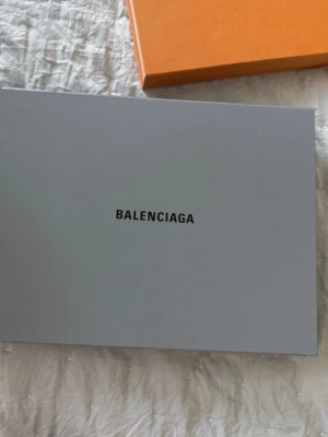 Balenciaga grå skokartong - Säljer en stilren grå skokartong från Balenciaga med svart logga på locket. Perfekt för förvaring eller som dekoration. Kartongen är rektangulär och tillverkad i hård kartong.kan gå ner i pris 