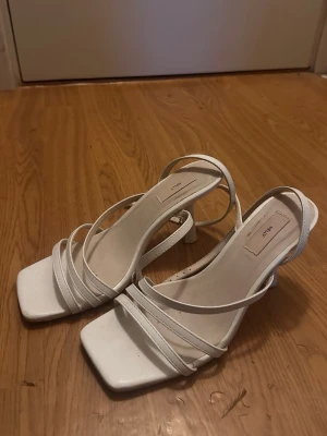 Vita sandaler med remmar från Nelly - Stilrena vita sandaler från Nelly med tunna remmar och fyrkantig tå. Skorna har en låg klack och öppen häl, vilket ger en trendig och modern look. Tillverkade i syntetmaterial och perfekta för sommarens outfits.