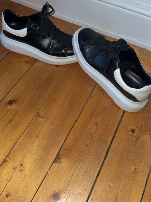 Svarta sneakers med vit sula - Säljer ett par svarta sneakers från Alexander McQueen med chunky vit sula och vit detalj vid hälen. Skorna har svarta skosnören och är tillverkade i läder för en clean och modern look. Perfekta för dig som gillar statement sneakers.