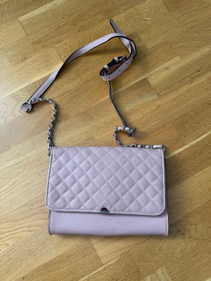 Ljusrosa quiltad handväska med kedja - Säljer en ljusrosa handväska med quiltat mönster på locket och en axelrem i kombinerat läder och kedja. Väskan har en stilren, rektangulär form och stängs med ett litet metallspänne framtill. Perfekt för dig som gillar trendiga accessoarer.
