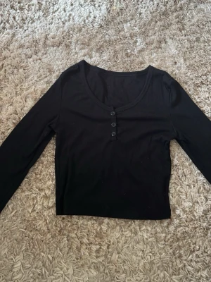 Svart långärmad topp med knappar - En enkel och stilren svart långärmad topp med rund halsringning och tre knappar framtill. Toppen är croppad och har en slim passform, perfekt att matcha med höga jeans eller kjol. Materialet känns mjukt och stretchigt.