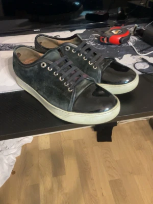 Svarta sneakers med lackdetaljer - Snygga svarta sneakers med glansiga lackdetaljer på tån och hälen. Ovandelen är i mockaliknande material och sulan är vit. Skorna har svarta skosnören och silverfärgade öljetter för en stilren look. Får ingen box med.