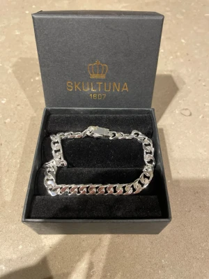 Silverfärgat kedjearmband Skultuna Nypris: 1000-1400 - Snyggt och stilrent armband från Skultuna i silverfärgad metall som är helt oanvänt. Nypris runt 1000-1400 kr. Armbandet har en chunky kedjedesign och kommer i originalask. Perfekt accessoar för att lyfta din outfit med en modern touch.