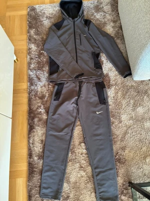 Grå Nike träningsset med huva - Snyggt grått träningsset från Nike med matchande hoodie och byxor. Hoodien har dragkedja, huva och svarta detaljer på axlar och ärmar. Byxorna har resår i midjan, svarta fickor och Nike-logga på benet. Perfekt för gymmet eller chill.