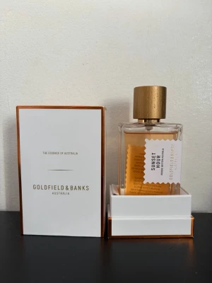 Goldfield & Banks Sunset Hour parfym - Exklusiv parfym från Goldfield & Banks i doften Sunset Hour. Flaskan är rektangulär med transparent glas och har en elegant guldfärgad kork. Parfymen har en varm bärnstensfärgad vätska och kommer i en stilren vit förpackning med gulddetaljer.