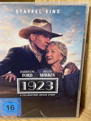 1923 Yellowstone  DVD-box - DVD-box med första säsongen av serien '1923', en Yellowstone Origin Story. Omslaget är i färg med westernmotiv och visar karaktärer i klassiska cowboykläder. Text på tyska och engelska. Innehåller bonusmaterial och är i plastfodral.