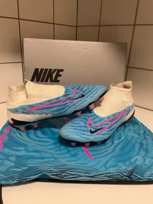 Nike blåa Phantom fotbollsskor - Snygga Nike Mercurial fotbollsskor i blått och vitt med rosa och svarta detaljer. Skorna har strumpliknande skaft och en modern design med mönstrad ovandel. Sulan är vit med rosa dobbar och blå linjer för extra grepp på planen. Perfekta för dig som vill sticka ut på fotbollsplanen. De är använda 1 gång och köpta från unisport. 
