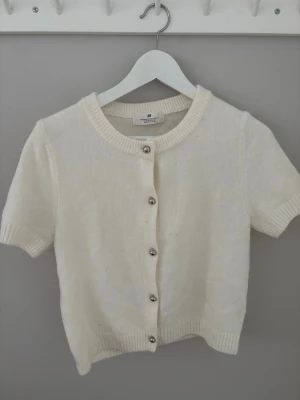 Vit kortärmad kofta från H&M - Söt vit kofta från H&M med korta ärmar och rund halsringning. Koftan har ribbade muddar och knäpps med silvriga knappar framtill. Perfekt att slänga över en topp för en enkel och clean look.