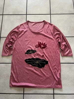 Rosa topp med blommigt motiv storlek L - Rosa topp med trekvartsärm och rund halsringning. Framsidan har ett broderat motiv med en mörkröd blomma och svarta blad. Materialet känns mjukt och stretchigt, troligen polyester eller bomullsmix. Perfekt för dig som gillar unika detaljer och blommiga inslag.