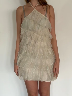 Glittrig volangklänning med halterneck - Supersöt kort klänning i ljus champagnefärg med glittrigt tyg och flera lager volanger. Halterneck med tunna axelband och öppen rygg ger en unik look. Perfekt för dig som vill sticka ut! Materialet känns lätt och skimrande.