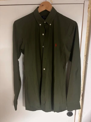 Olivgrön skjorta från Polo Ralph Lauren - Snygg olivgrön skjorta från Polo Ralph Lauren med klassisk krage och knäppning framtill. Skjortan har långa ärmar och den ikoniska röda logotypen broderad på bröstet. Perfekt till jeans eller chinos för en clean look. Använd fåtal gånger, köpt på zalando för 900kr