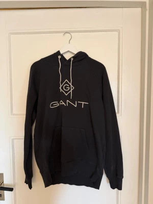 Svart hoodie från GANT med logga - Säljer en svart hoodie från GANT med stor vit logga och emblem på bröstet. Hoodien har huva med vita snören och en stor magficka framtill. Tillverkad i mjukt material och har ribbade muddar vid ärmar och nederkant. Perfekt för en avslappnad och stilren look.