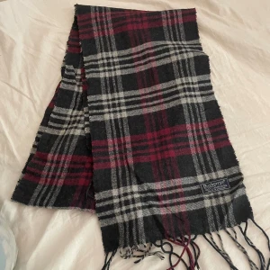 Rutig halsduk från Burberrys i ull - Snygg rutig halsduk från Burberrys i 100% lammull. Färgkombinationen är svart, grå och vinröd med klassiskt rutmönster och fransar i ändarna. Perfekt accessoar för att lyfta din vinterstil.