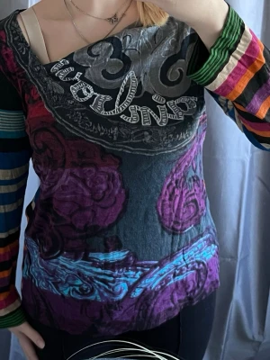 Färgglad randig topp från Desigual - Säljer en unik topp från Desigual med färgglada ränder på ärmar och rygg samt tryck i lila, blått, grått och svart framtill. Toppen har lång ärm, normal passform och är tillverkad i mjukt material.  Toppen är i väldigt fint skick utan några fläckar eller hål.