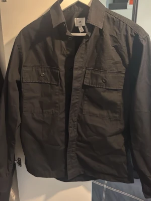 Svart skjorta från H&M med fickor - Stilren svart skjorta från H&M med två stora bröstfickor och klassisk krage. Skjortan har långa ärmar och knäppning framtill. Perfekt för en clean och enkel look. Använts 2-3 gånger. 
