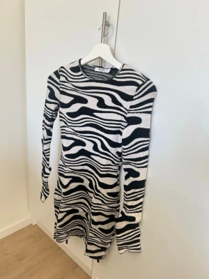 Zebramönstrad stickad klänning - Säljer en zebramönstrad stickad klänning från NA-KD i svart och vit. Klänningen har lång ärm, kort längd och en stor öppen rygg med snörningsdetaljer. Perfekt för dig som vill sticka ut med ett djurmönster och cool design.