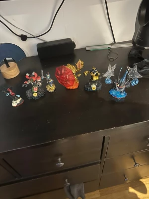 Skylanders figurer till spelkonsol - Säljer flera Skylanders-figurer i olika färger och former, perfekta för användning med Skylanders-spel på konsol. Figurerna är tillverkade i plast och har detaljerade designer med bland annat blå, röd, gul och svart färg samt genomskinliga och metalliska inslag.
