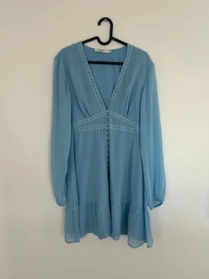 Ljusblå klänning med detaljer - Supersöt ljusblå kortklänning från NA-KD med v-ringning, detaljer och knappar framtill. Klänningen har långa, luftiga ärmar och volang nertill. Perfekt för vår och sommar!